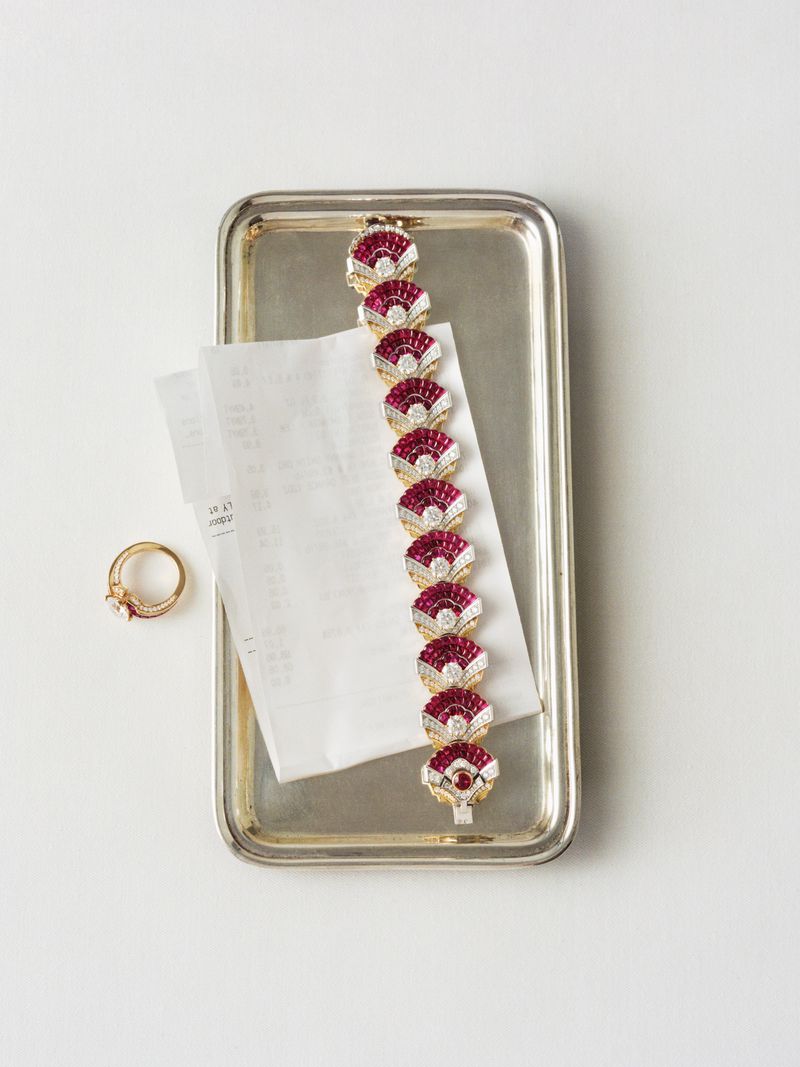 Overhead shot of a ruby and diamond bracelet, a silver tray, a ring, and a receipt on a white surface, product photography.