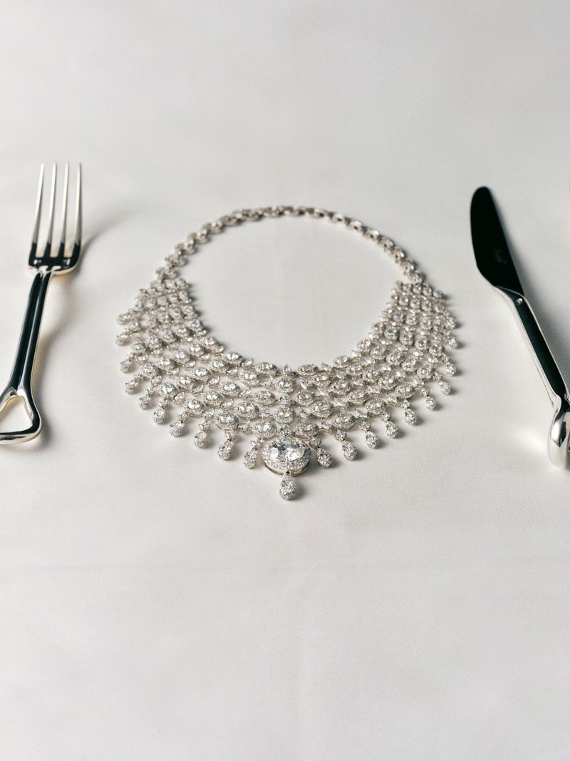 Diamond necklace arranged on a white tablecloth between a fork and knife, presented as part of a luxurious, jewelry-themed table setting.