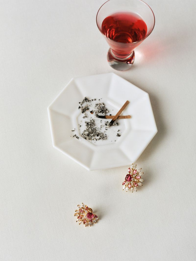 Close-up still life shot features a cocktail glass, ashtray with cigarette butts, and jewelry on a clean, white background.