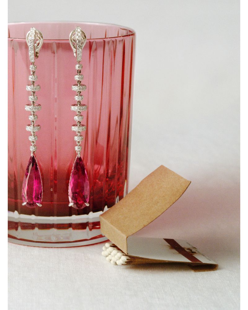 Elegant long drop earrings with pink gemstones and diamonds displayed next to a pink glass and small box on a white surface.