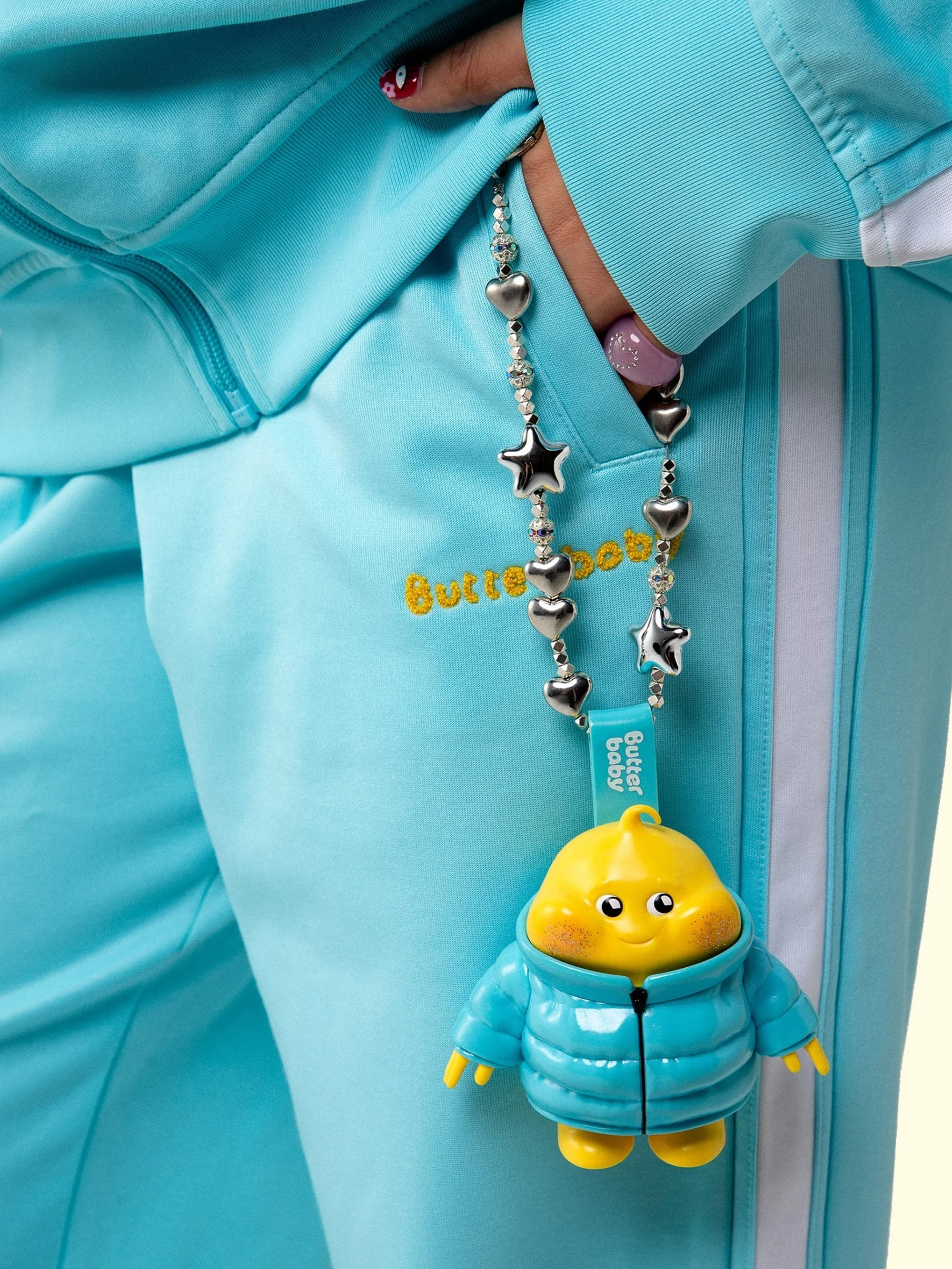 Close-up of a blue tracksuit with yellow embroidered logo and a 'Butter Baby' keychain featuring a yellow doll wearing a blue jacket.