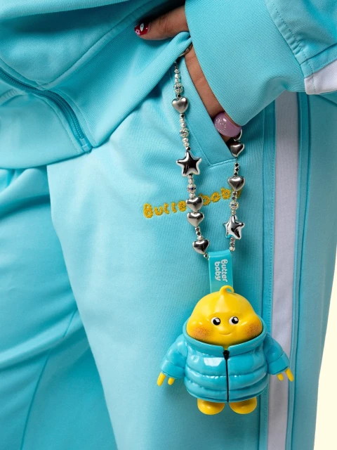 Close-up of a blue tracksuit with yellow embroidered logo and a 'Butter Baby' keychain featuring a yellow doll wearing a blue jacket.