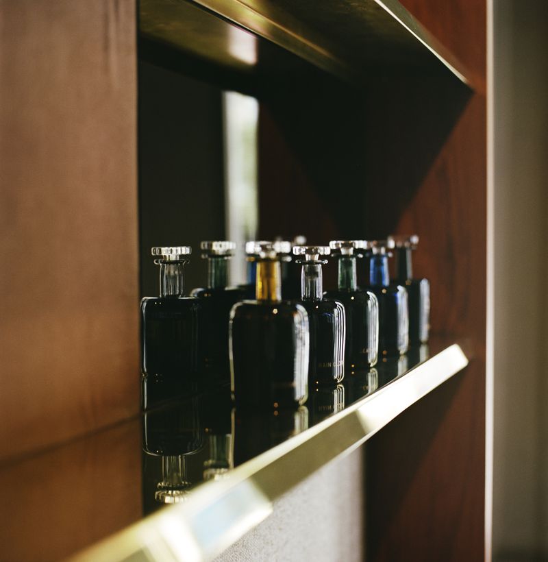 Row of dark glass bottles with silver tops sits on a reflective shelf against a brown panel, conveying a sense of luxury and refinement.