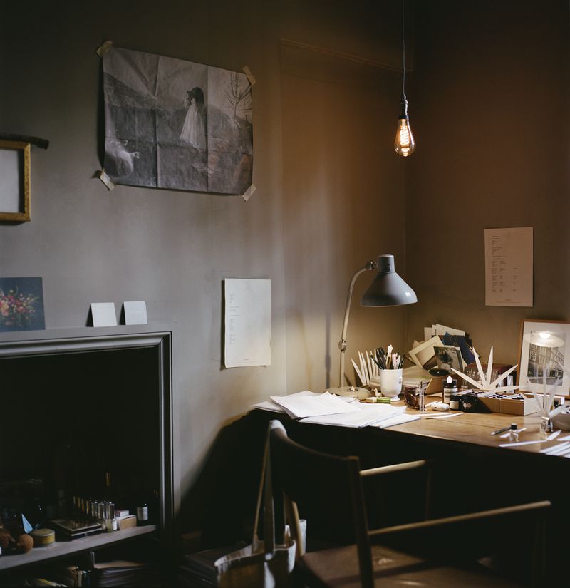 Interior scene shows a desk with stationery, a lamp, and a chair in a room with neutral walls and warm lighting from an overhead bulb.
