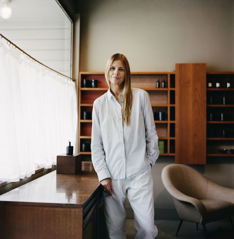 Blond woman in light-grey shirt and pants stands in interior with wooden shelves and modernist furniture; indoor setting with natural light.