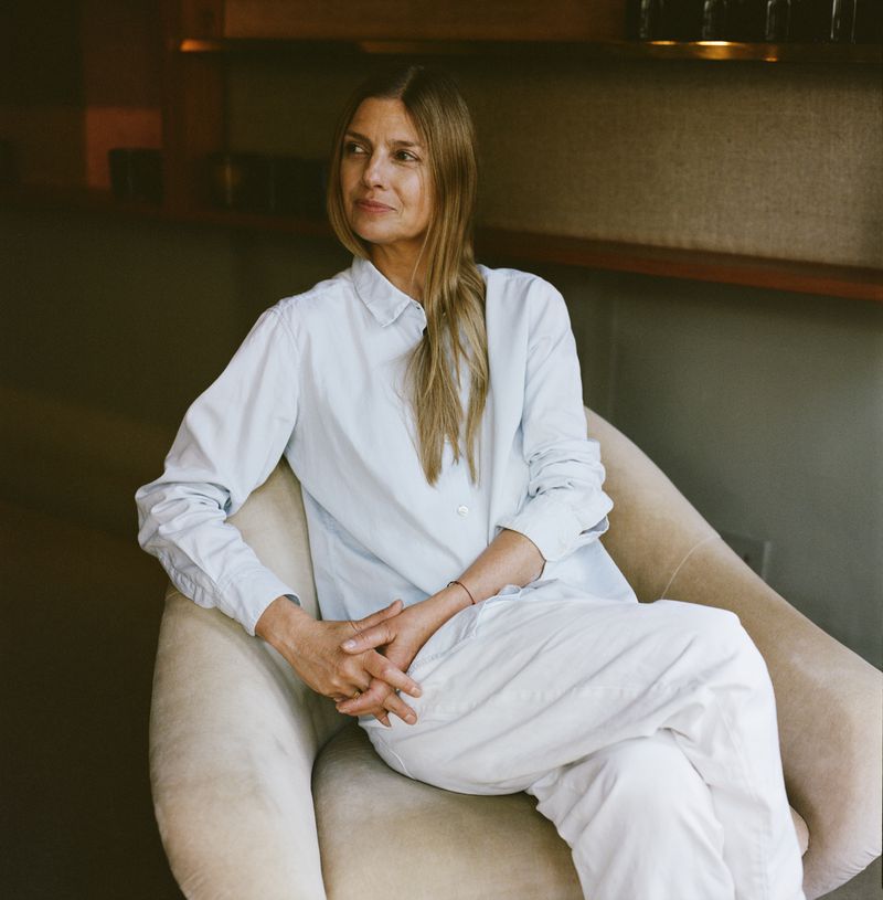 Portrait of an adult woman in a button-down shirt and white pants sitting in a tan chair in a relaxed pose indoors.