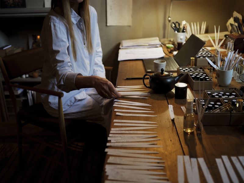 Adult woman sits at a wood table with many white strips in a dimly lit room with laptop, tea pot, tea cup, and vials.