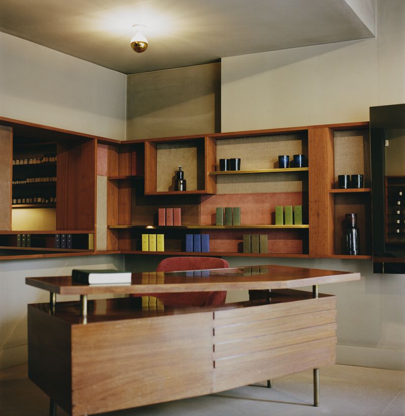 Interior view of a modern home office featuring a wooden desk, built-in shelving filled with books and decorative objects, and warm lighting.