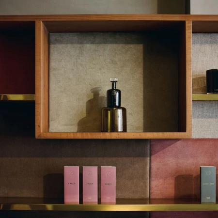 Still life shot of a glass perfume bottle on a shelf, accompanied by pink-packaged beauty products and a wooden, multi-sectioned display case.
