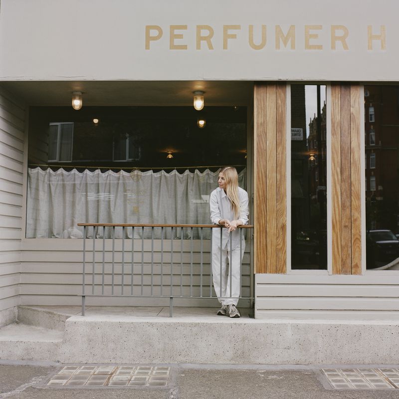 Blonde woman stands in front of the Perfumer H store with a light gray exterior, wooden trim, and a metal railing outdoors.