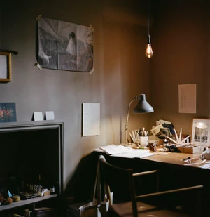 An artist desk with papers, lamp, and stool against a grey wall in a warm interior room.