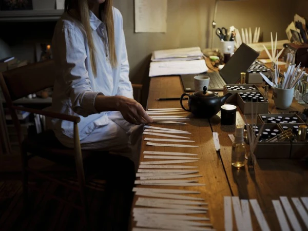 Woman in a white shirt sitting at a wooden workbench lined with paper blotters and perfume bottles