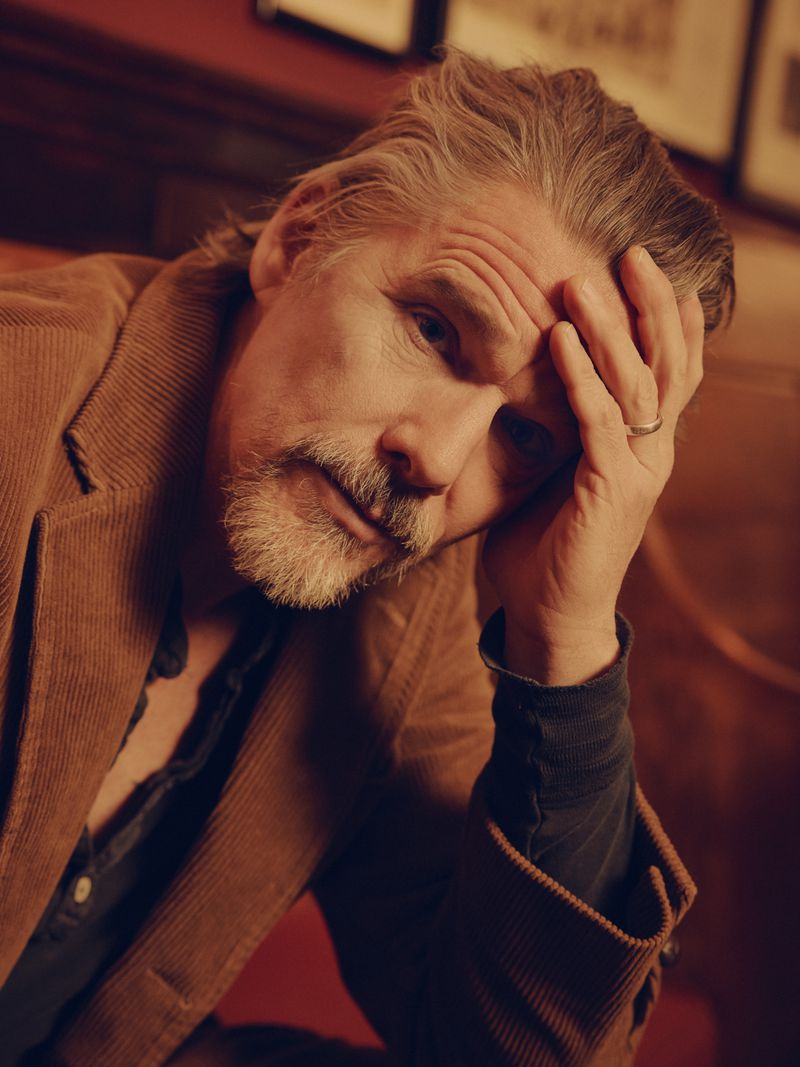 Portrait of a grey-haired man in a corduroy jacket with his hand on his head, looking thoughtfully into the camera indoors.