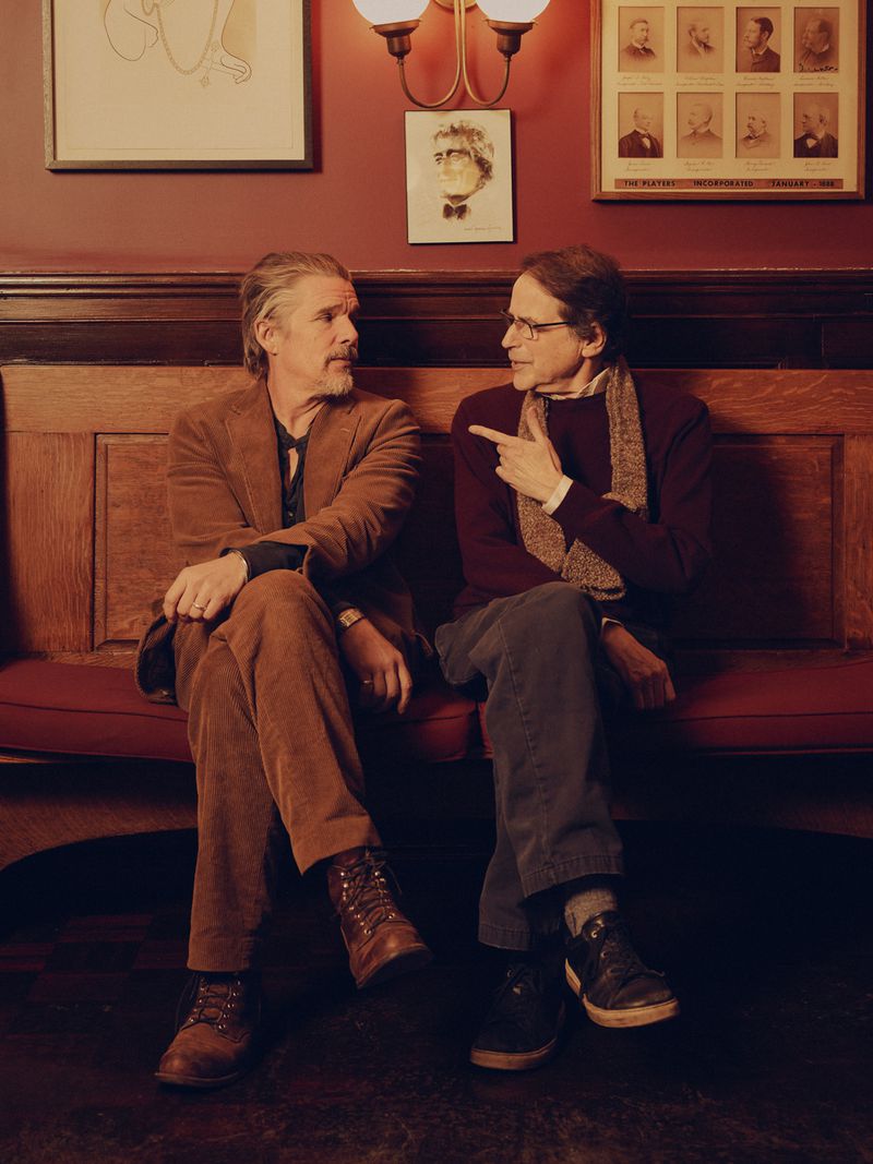 Two men seated indoors, one in a brown corduroy suit and the other in a sweater, conversing beneath vintage-style framed artwork and sconces.