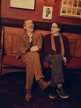 Two adult males sitting closely on a bench in an indoor setting, one is laughing.  Dark wood details and framed photos are visible in the background.