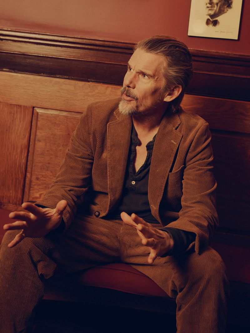 Adult male in a corduroy suit sits indoors, looking off-camera with open hands. Portrait of a man in a formal setting.