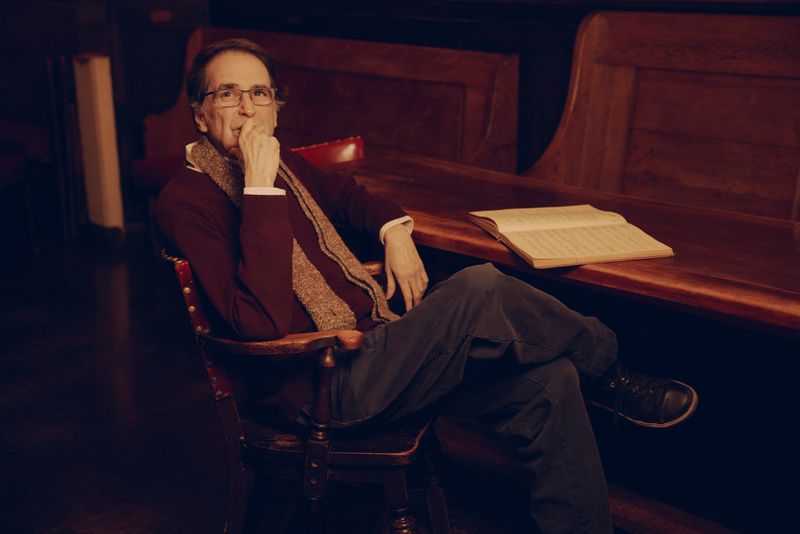 Man in glasses sits in a wooden chair inside a wood-paneled room next to a wooden table with a book; dark moody lighting.