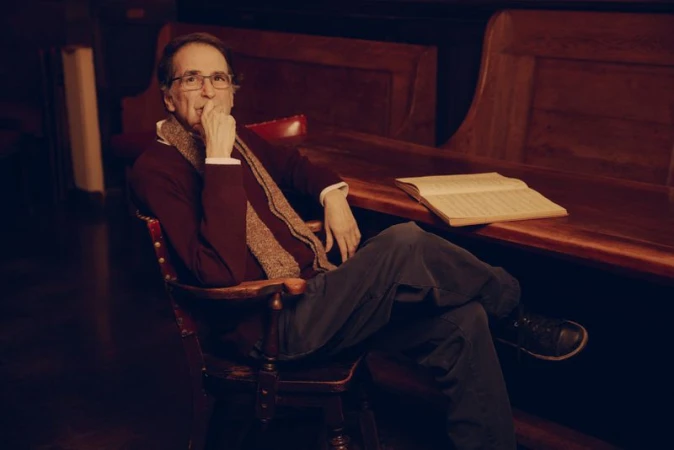 Man in glasses sits in a wooden chair inside a wood-paneled room next to a wooden table with a book; dark moody lighting.