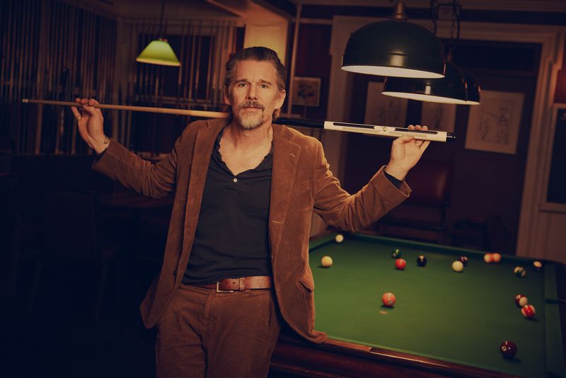 Ethan Hawke poses at a billiards table in a corduroy suit, holding a pool cue. The setting features billiards equipment in a warm, subdued light.