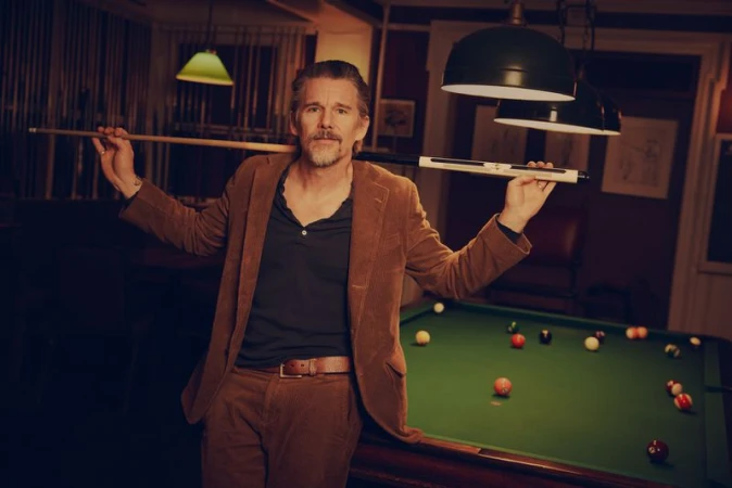 Ethan Hawke poses at a billiards table in a corduroy suit, holding a pool cue. The setting features billiards equipment in a warm, subdued light.