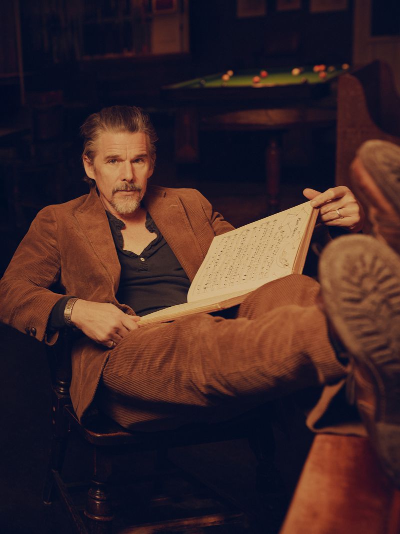 Adult male in a brown corduroy suit sits in a chair, holding a book with musical scores, pool table in background, warm lighting.