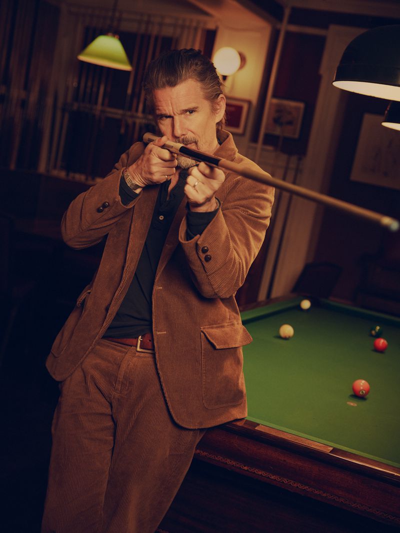 Mature adult male wearing a tan corduroy suit poses with a pool cue at a billiard table in an indoor setting with warm lighting.