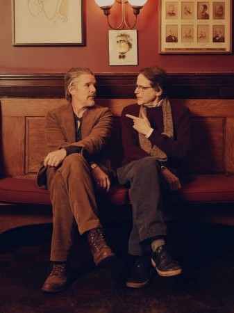 Ethan Hawke and Robert Kaplow seated together on a wooden bench against a red wall for LA Times by photographer David Urbanke.