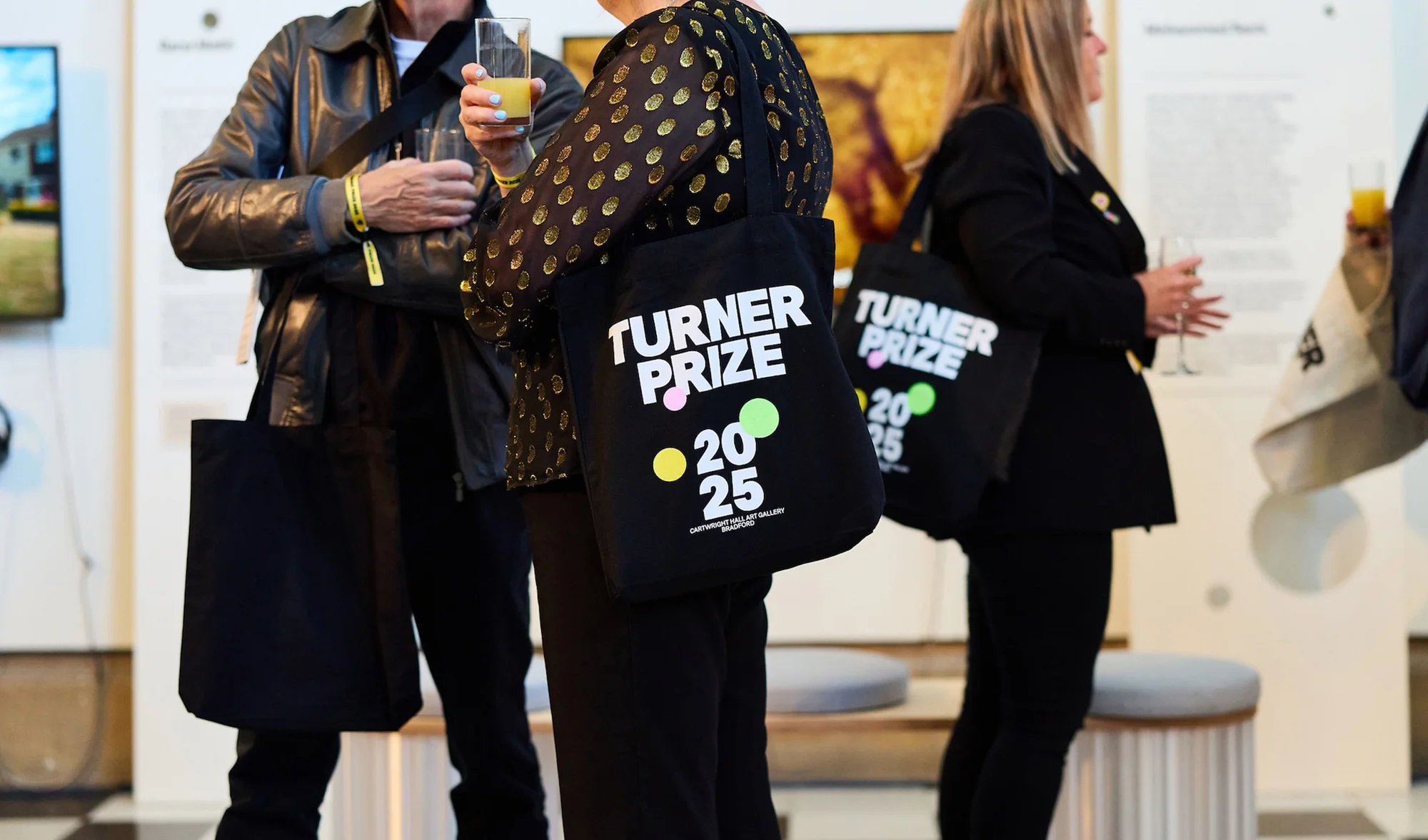 Attendees at the Turner Prize 2025 event carrying branded tote bags, dressed in black, holding drinks, and standing inside an art gallery.