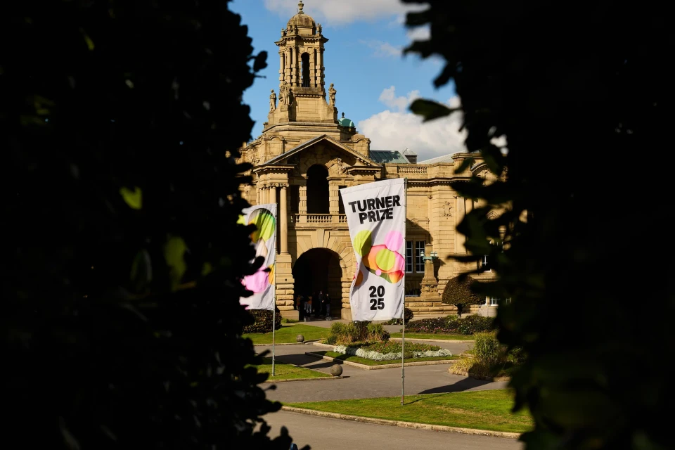 Exterior view of a grand building framed by dark foliage, with 'Turner Prize 2025' banners displayed on the front lawn.