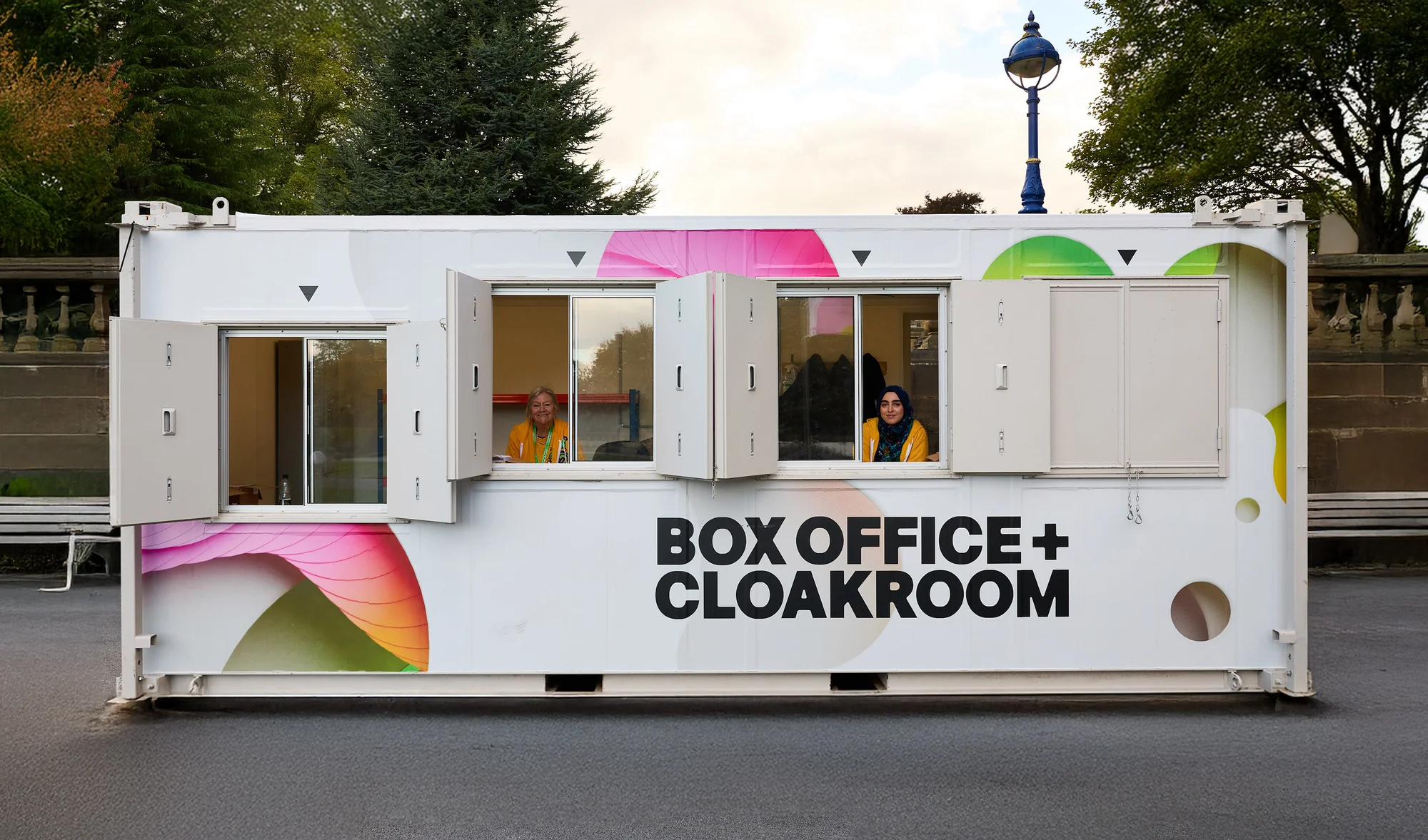 Outdoor shot of a white portable box office with two open window counters; two people are visible inside each wearing yellow shirts.