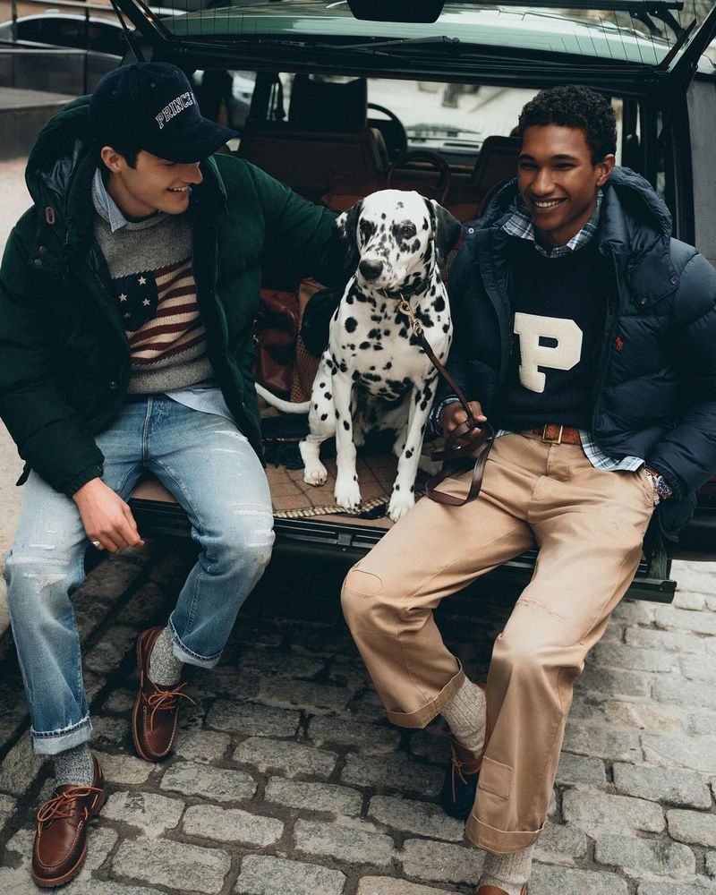 Two adult men and a dalmatian dog sit in the open trunk of an SUV wearing preppy sweaters, jackets, pants, shoes on a cobblestone street.
