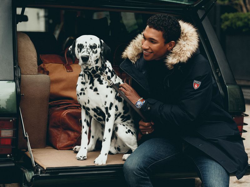 Man with a Dalmatian dog sitting in the back of a car filled with luggage. He is wearing a fur-lined jacket and jeans.