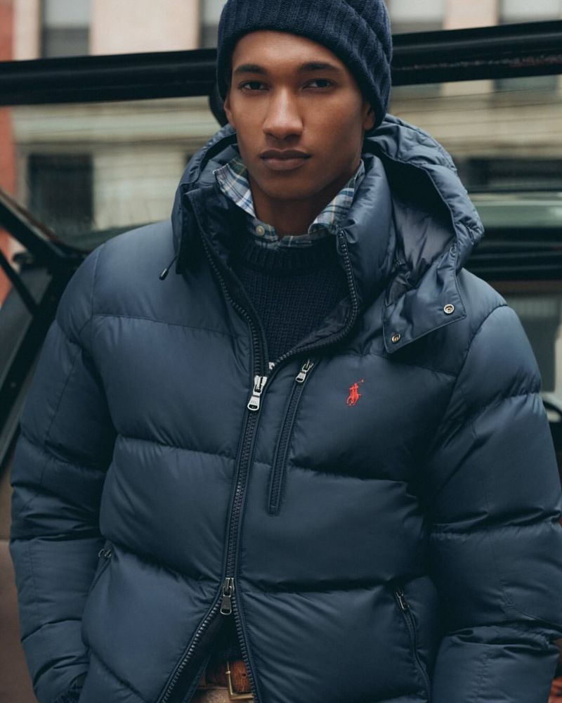 A man in a navy blue puffer jacket and beanie stands outdoors in an urban setting, with a building visible in the background.