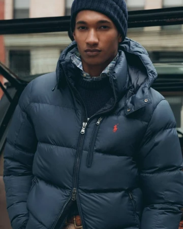 A man in a navy blue puffer jacket and beanie stands outdoors in an urban setting, with a building visible in the background.