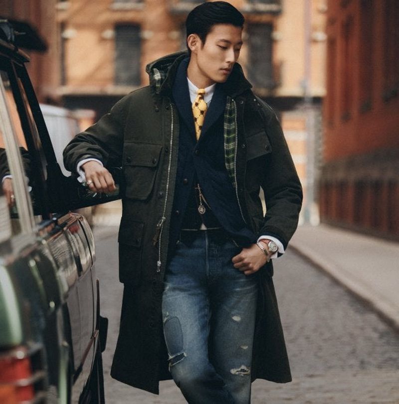 An adult male in a stylish olive green coat and blue jeans stands next to a dark SUV in an urban setting, exuding a fashionable vibe.