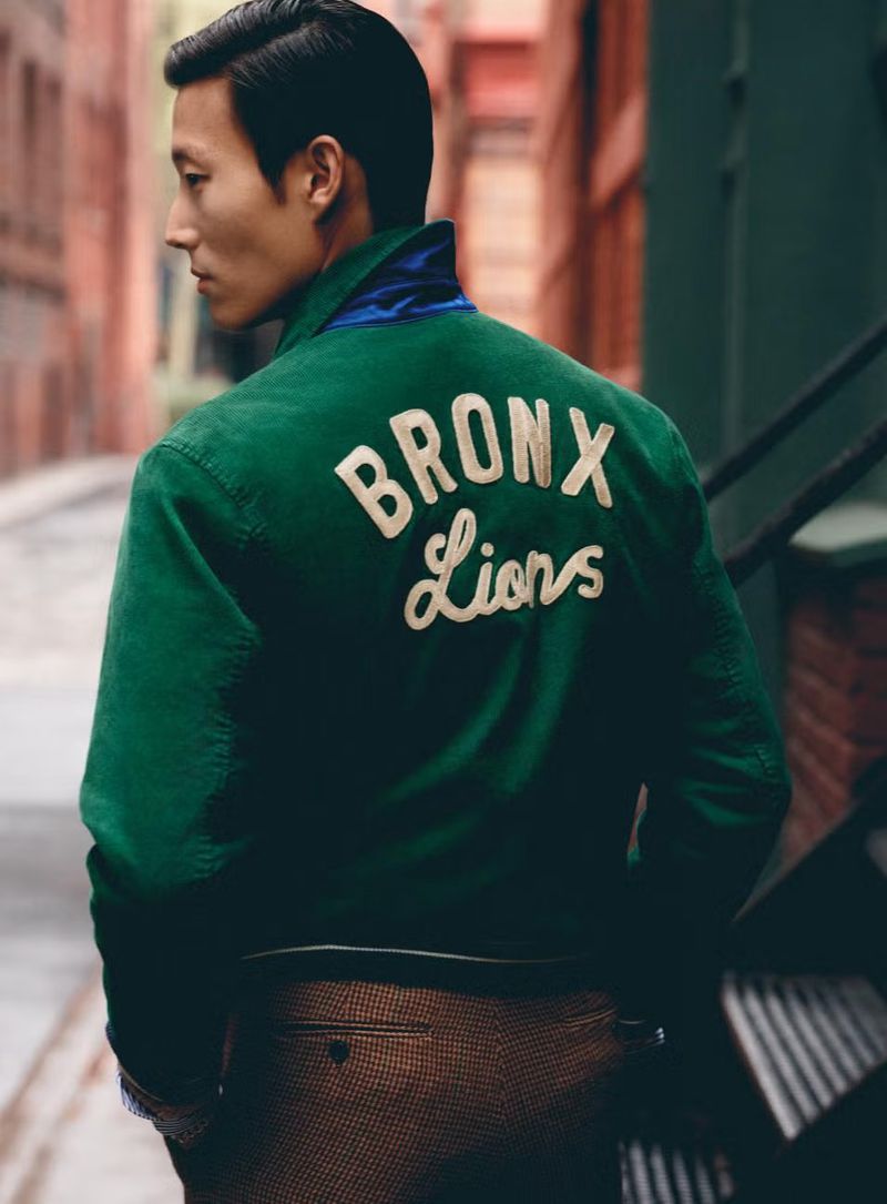 Adult male poses in a fashion shot outdoors wearing a green jacket with 'Bronx Lions' on the back; he wears brown pants.