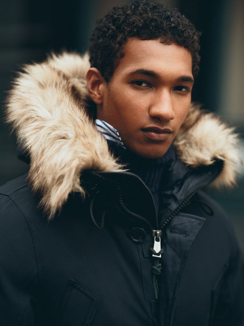Portrait of a stylish adult male in a navy blue parka with a fur-trimmed hood, looking directly at the camera with a neutral expression.