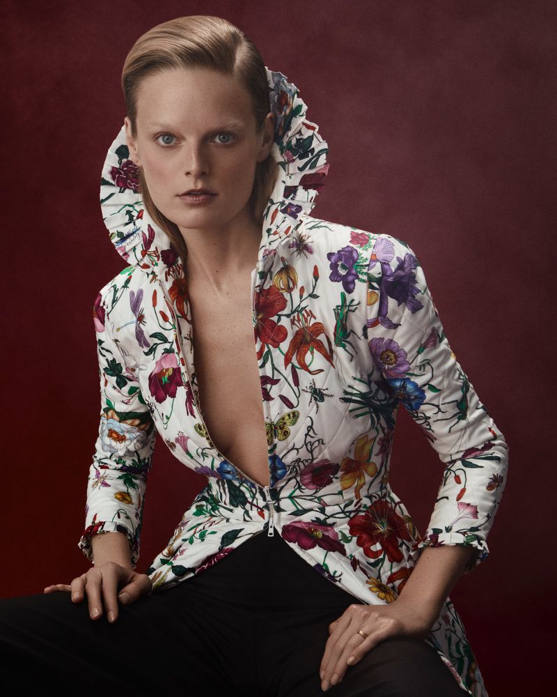 Fashionable adult woman in floral jacket poses in studio setting with simple red background, highlighting style and design details.