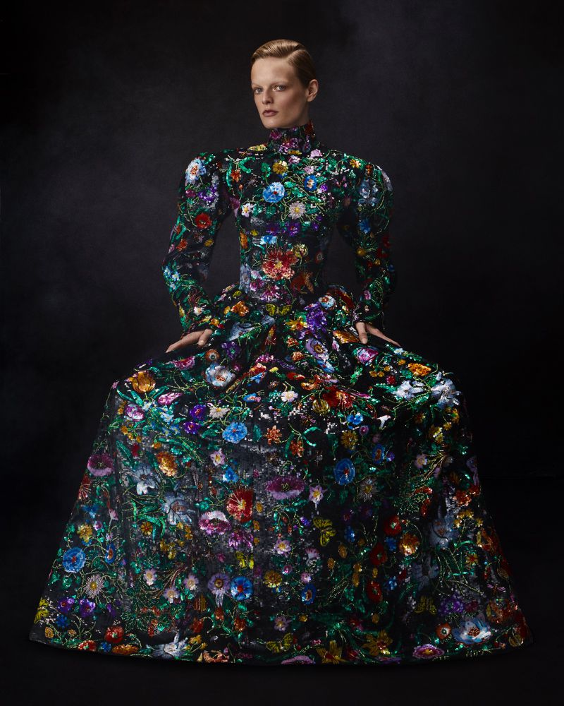 A fair-skinned woman in a colorful floral gown poses against a dark, smoky backdrop in a fashion-forward, eye-level portrait.