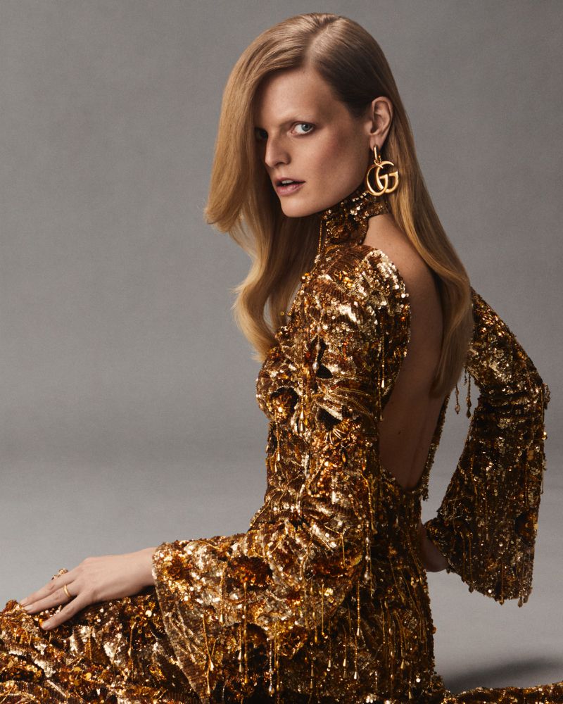 Woman with long blonde hair sits in a gold sequined dress and matching earrings against a grey background in a fashion advertisement.