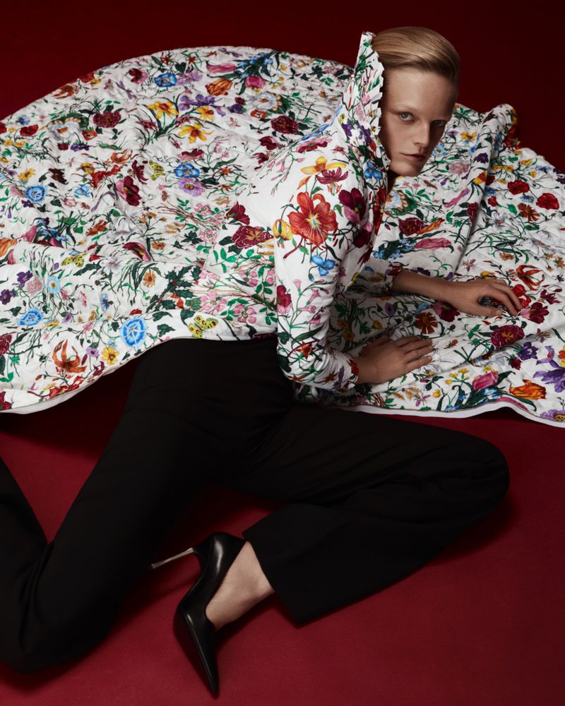 Fashion shot of a woman in a floral coat and black pants lying on a red surface, staring intently at the camera with a neutral expression.