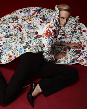 Fashion shot of a woman in a floral coat and black pants lying on a red surface, staring intently at the camera with a neutral expression.