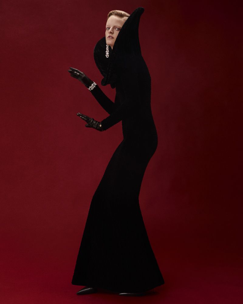 A person in a black gown with a high collar poses against a red background, accessorized with jewelry and gloves for a fashion editorial.