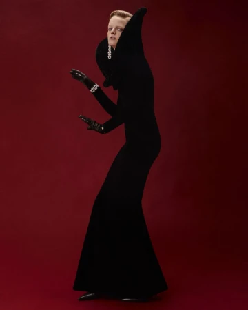A person in a black gown with a high collar poses against a red background, accessorized with jewelry and gloves for a fashion editorial.