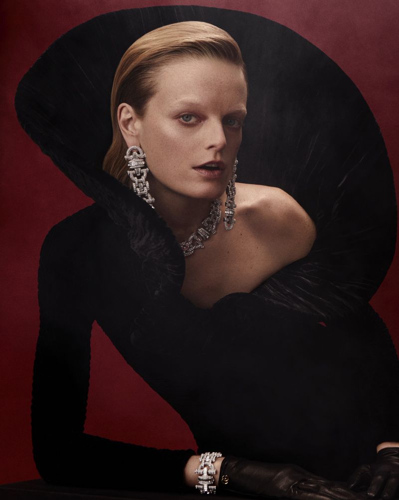 A model with blond hair is posing indoors wearing a black dress, diamond earrings, and necklace against a red studio background.