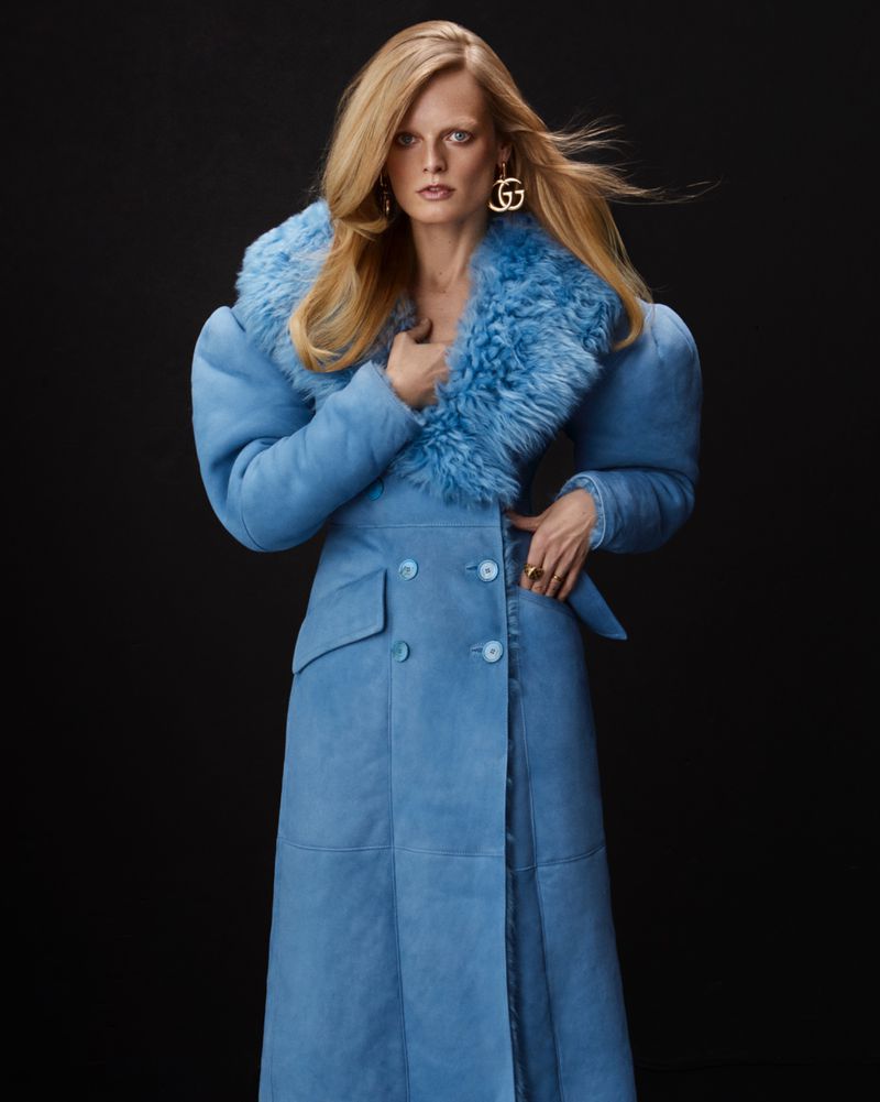 A blonde-haired woman poses in a long, light-blue shearling coat and gold logo earrings against a black background in a studio shot.