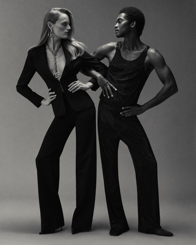 Monochrome studio shot of a blond female model in a black suit, standing next to a black male model in a sparkling black top and pants.