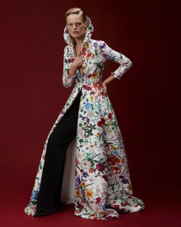A fashion model poses in a white floral coat and black trousers against a red background, looking directly at the viewer.