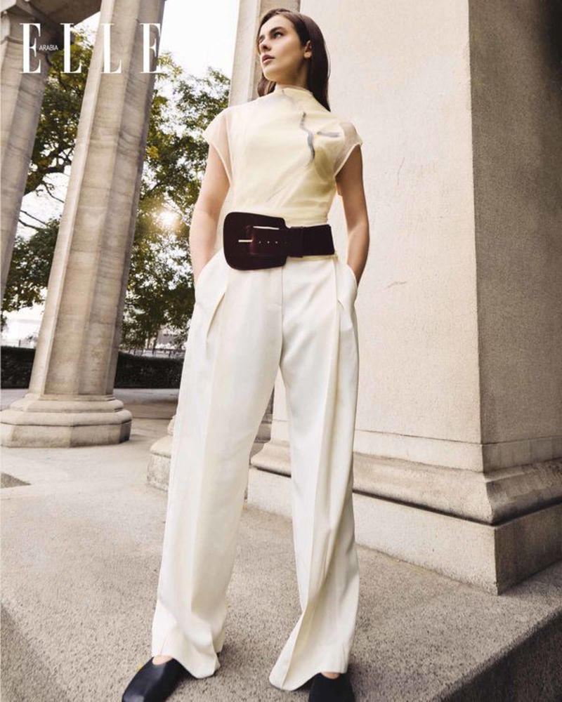 Fashion editorial shot of a woman in white pants and a cream-colored top posing outdoors with columns in the background, looking away.