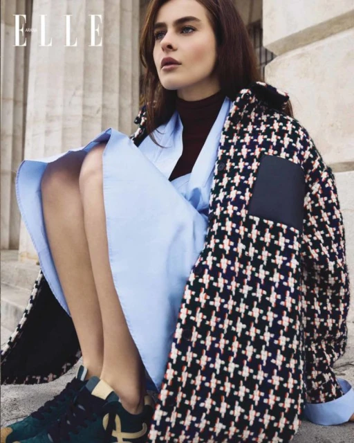 Fashion editorial shot of a young woman sitting on stone steps wearing a blue dress and patterned coat with sneakers on steps.